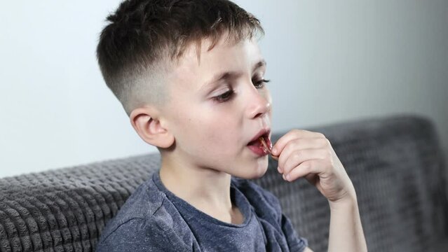 A little boy opening sweets and taking pastila his mouth while sitting on a cozy sofa. A child enjoys candy