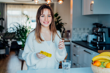 Young smiling woman holding pills organizer at home kitchen. Positive lady takes daily medicine...