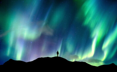 Aurora borealis glowing over silhouette hiker standing on the mountain in the night sky