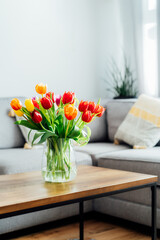 Vase of fresh tulips on the coffee table with blurred background of modern cozy light living room with gray couch sofa, graphic cushions and green plants. Open space home interior design. Copy space © okrasiuk