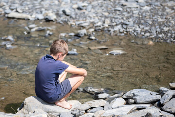 Little boy sitting on rocks with his back looking at the mountain lake.