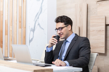 Handsome serious businessman in black suit sitting at the desk and working on laptop in the modern office	
