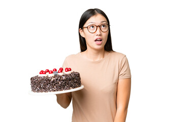 Young Asian woman holding birthday cake over isolated chroma key background looking up and with surprised expression