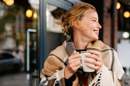 Joyful blonde woman in warm plaid drinking hot tea while stanging outdoors