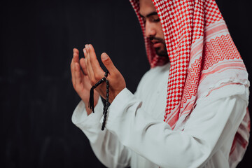 Arabian man in traditional clothes making traditional prayer to God, keeps hands in praying gesture in front of black chalkboard representing modern islam fashion and ramadan kareem concept