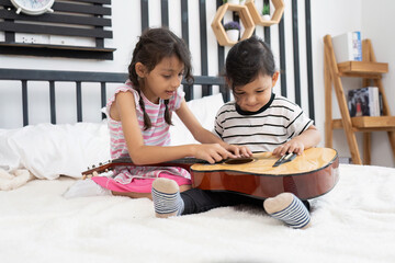 Fototapeta premium Cute small boy and his sister sitting try to play guitar on bed in the bedroom