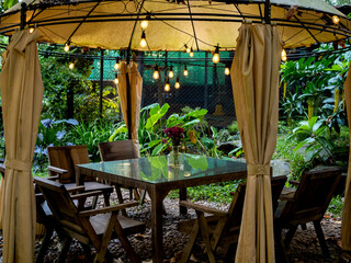 Vintage light bulbs hanging on the old tent over the empty dining table set, glass table and wooden chairs in the outdoor garden, evening scene with no people.