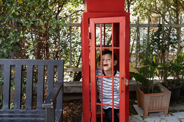 Little boy laugh playing inside a public red phone booth look outside to parent