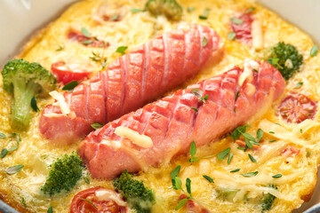 Omelette or frittata with two sausages, melted cheese, cherry tomatoes, broccoli and parsley on red skillet or iron pan on white old concrete background. Healthy breakfast food concept. Top view.