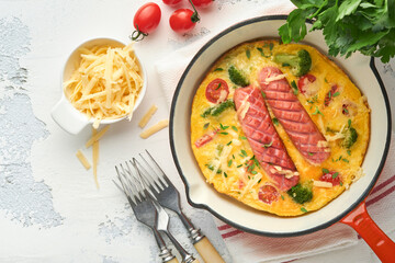 Omelette or frittata with two sausages, melted cheese, cherry tomatoes, broccoli and parsley on red skillet or iron pan on white old concrete background. Healthy breakfast food concept. Top view.