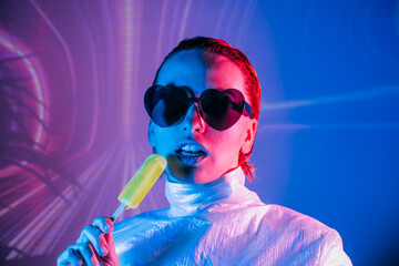 Portrait of a cyberpunk girl in heart shaped sunglasses in neon light. Seductive woman licking a...