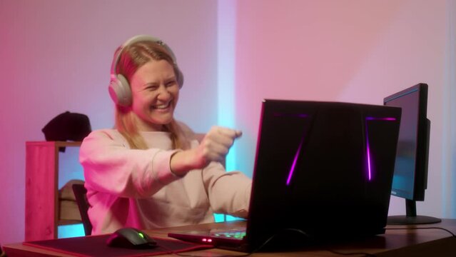 girl gamer plays on computer in purple-blue backlight of room and wins online match in game. young woman rejoices and raises her hands up in happiness. Winning difficult deathmatch.