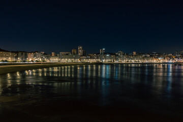 Naklejka na ściany i meble The city is illuminated at night. Embankment near the sea, the rays are reflected in the water.