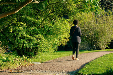 Black woman walks in a park. Unrecognizable woman with afro hair strolls along a tree-lined avenue during sunset. Image of serenity on an autumn day. Back view of womanin black dress with shoulder bag