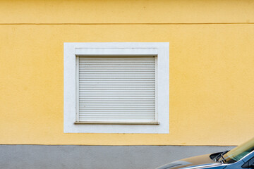 The window in the house is covered with white shutters on a yellow wall.