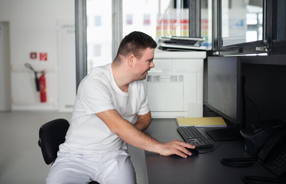Young Man With Down Syndrome Working In Hospital Office, Writing Something On Computer. Concept Of Integration People With Disabilities Into Society.