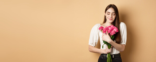 Valentines day. Tender and romantic girl, smell roses and smile with closed eyes. Girlfriend hugging gift flowers from lover, standing on beige background