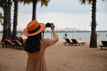 A middle-aged female tourist uses a small digital camera to capture the splendor of the sea in the dim evening. A woman wearing a light brown dress, an orange wide-brimmed hat is happy to take picture