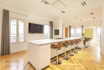 Large living room with varnished hardwood floors with a long white high top table with many tall wooden and chrome metal stools