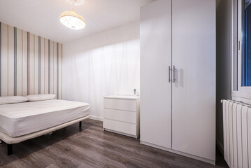 Bedroom with white wooden wardrobe, chest of three drawers and undressed bed