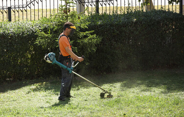 A worker mows grass with a mechanical trimmer. Improvement of a green lawn with a manual lawn mower with an internal combustion engine. Work with landscape design