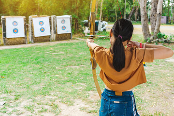 Back view of Asian girl wear face mask aims archery bow and arrow to colorful target in shooting...