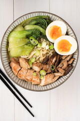 Asian noodle soup, ramen with chicken breast, vegetables, mushrooms, egg and chopsticks on a white wooden table. Top view, healthy eating concept