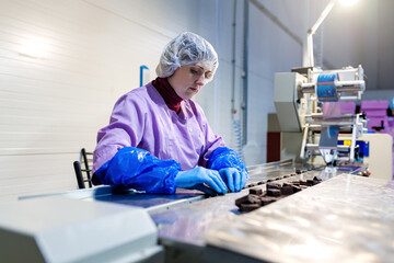 Professional female worker in uniform and protective gloves sorts chocolate candies on production line at factory - 563967258