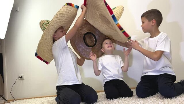 three boys brothers mexican cowboy hats take off each other try on different hat on little boy brother wear biggest headdress mexican hats white t-shirts backlit room white carpet friendship travel