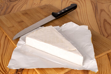 One slice of delicious brie cheese with wooden kitchen board and metal knife on wooden table, macro.