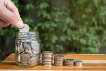 hand of a man saving coin in a jar, with natural lighting and coin on wooden table with green background, symbol of money saving for retirement