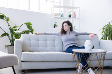 Fototapeta premium Young woman sits relaxed on a sofa in her living room with her arms spread to both sides looking over her shoulder to the right with a happy expression on her face