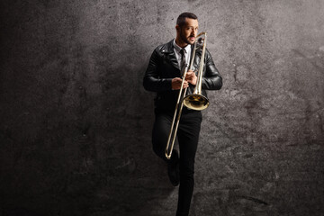 Man in a leather jacket holding a trombone and standing against a gray wall