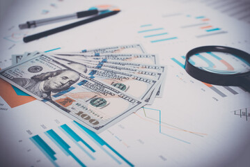 Close-up of money, business documents, magnifier, pen and pencil in workplace. Low angle view, selective focus on one hundred dollar bills