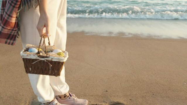 A girl stands on the sand next to the sea waves and holds a basket of Eggs for Easter. I'm getting closer, slow motion.