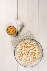 Glass bowl with popcorn, popcorn kernels and salt on a white wooden table, top view, copy space