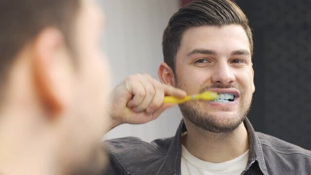 Handsome young man brushing his teeth looking at himself in the mirror, loves white teeth, enjoys personal care.