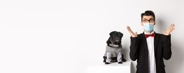 Coronavirus, pets and celebration concept. Amazed young man in face mask and suit staring at camera surprised, cute black dog sitting near owner in party outfit, white background
