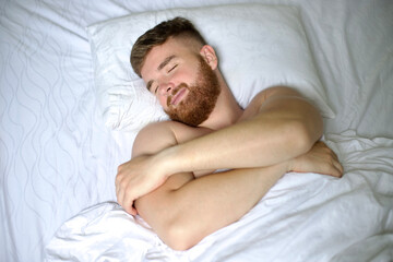 Handsome young calm sleepy bearded man with beard is sleeping well on side in bed in bedroom on pillow with his eyes closed in the morning, resting. Good healthy dream, rest. White linens. Top view.