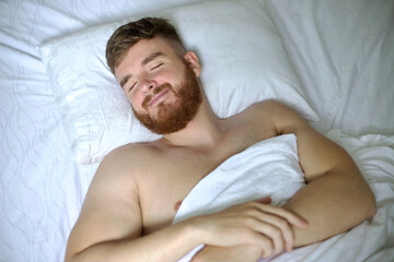 Handsome young calm sleepy bearded man with beard is sleeping well on side in bed in bedroom on pillow with his eyes closed in the morning, resting. Good healthy dream, rest. White linens. Top view.