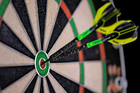 Two black and green darts stuck in the bullseye of a dartboard. Close up of a dartboard.