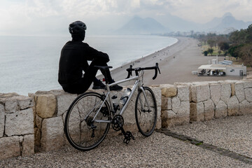 cycling composition. a man in a cycling suit sits with his back near a bicycle on a serpentine against the background of the sea. person riding a bicycle