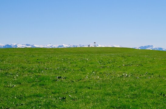Green meadow against blue sky, people, wayside cross and snowy mountain range on the horizon. Hochberg near Pfaender. Vorarlberg, Austria.