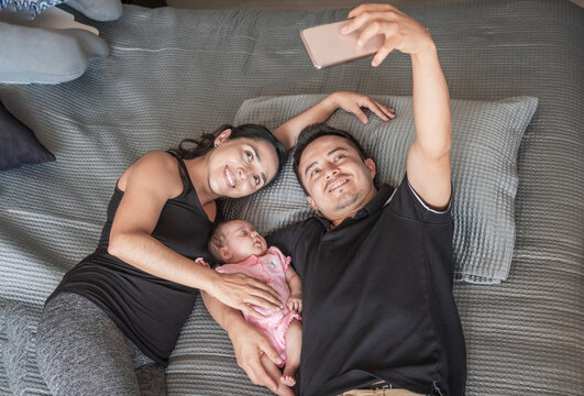 Smiling Father And Mother Taking A Selfie With Their Baby In Bed