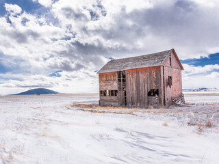 Red abandoned barn in an open field, San Luis Valley, Colorado, with fence, bright blue sky, sunshine, snow, and clouds