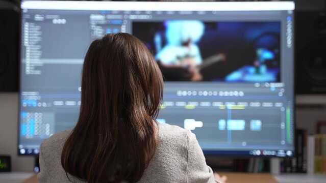 Back view of female video editor editing new project film montage on full screen computer sitting at office or home. Content creator in digital company processing movie with post production software