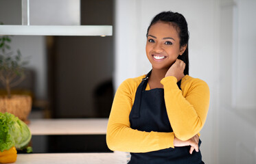Fototapeta premium Smiling young african american lady cook in apron near table with vegetables in minimalist kitchen interior