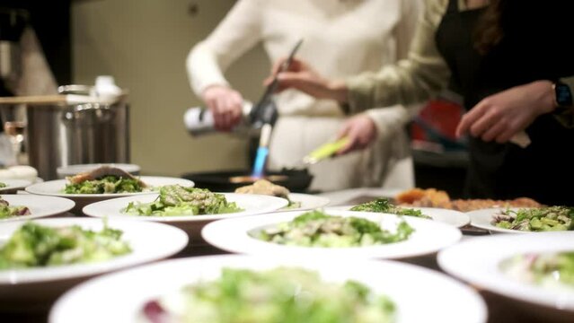 One chef roasts salmon with a gas burner. The second arranges the finished salmon on plates. In the foreground are plates of salad for a group of people. The concept of cooking dinner.