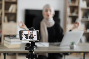Blurred relaxed young muslim woman in casual wear sitting on workplace with modern laptop and meditating with closed eyes during recording video blog. Focus on modern smartphone fixed on tripod.