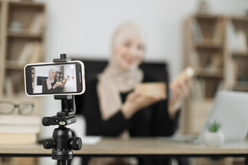 Focus on phone of happy muslim female blogger unpacking gift boxes presents from companies and doing live streaming. Arab woman using smart phone camera for creating content.
